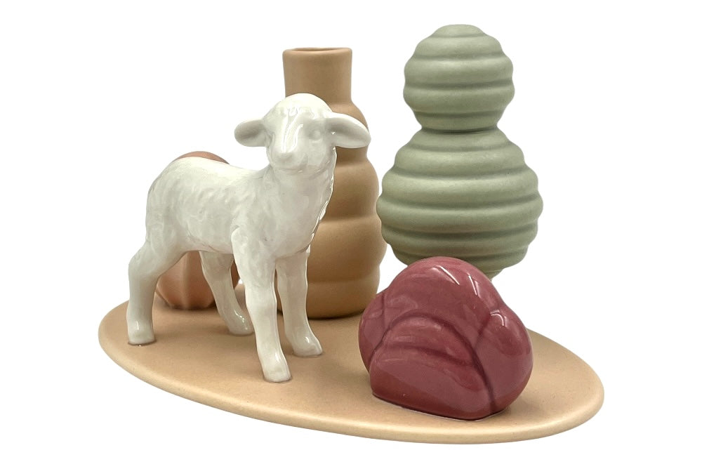 Dekorative Keramikvase mit einer weißen Lammfigur im Zentrum, umgeben von abstrakten Keramikelementen in erdigen und pastelligen Farben. Dazu gehören eine beige Vase, eine grüne Vase und Deko-Elemente in Rosé und Dunkelrot. Alles steht auf einer ovalen Sandtonplatte.